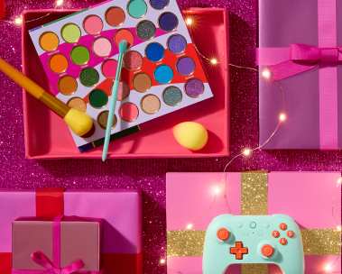 A colorful assortment of gifts on a pink surface, featuring a game controller and makeup items.