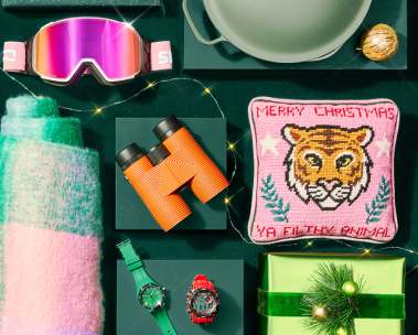 Dunkelgrüner Hintergrund. Sortiment an weihnachtlichen Geschenkartikeln (rosa Kissen, Skibrille, Uhr, Schal, Pakete)